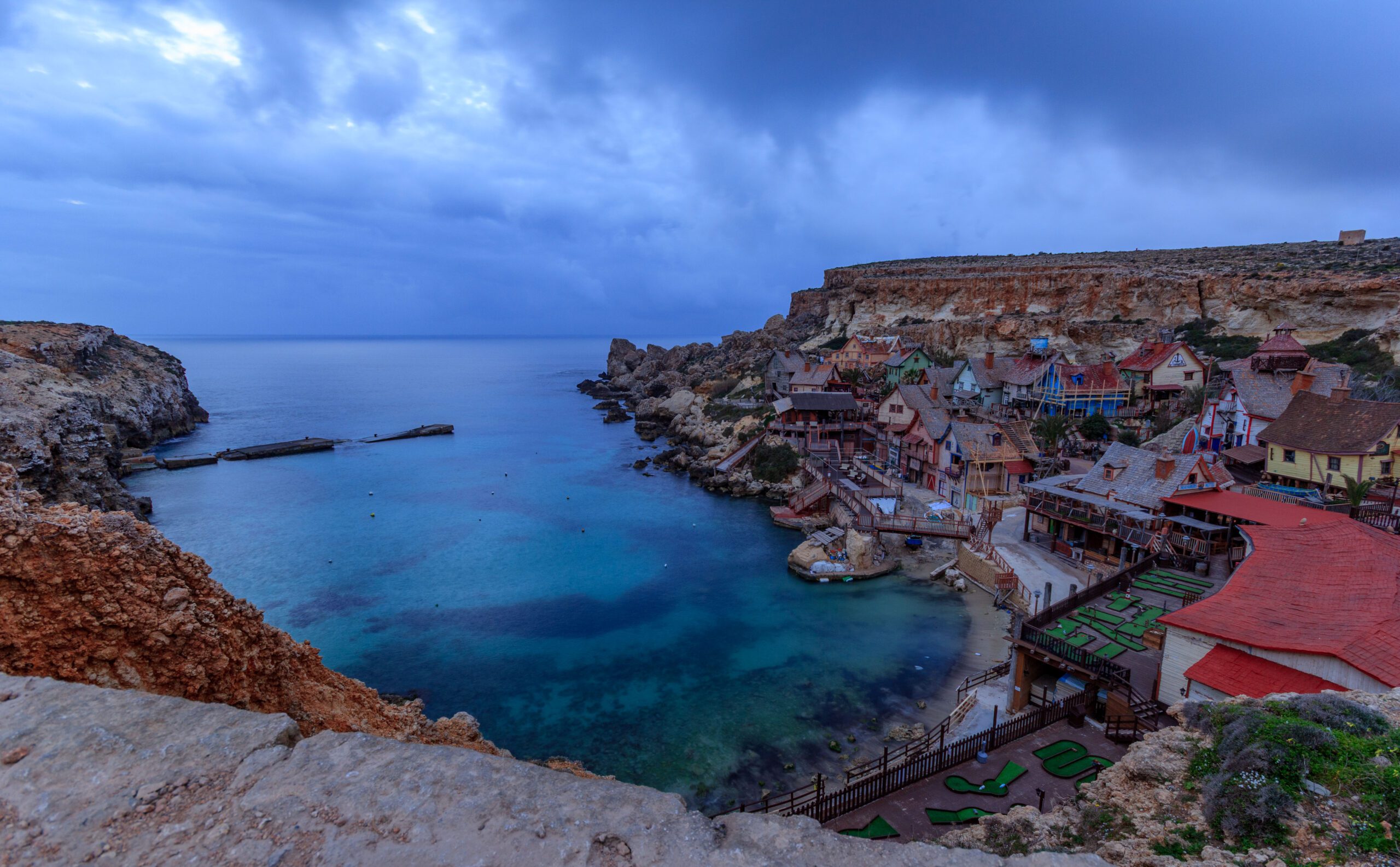 Popeye Village in Malta during blue hour on a cloudy day, with colourful wooden houses along the coastline