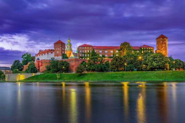 Wawel Royal Castle in Kraków, Poland, during blue hour with lights from the castle and walking path reflected in the Vistula River