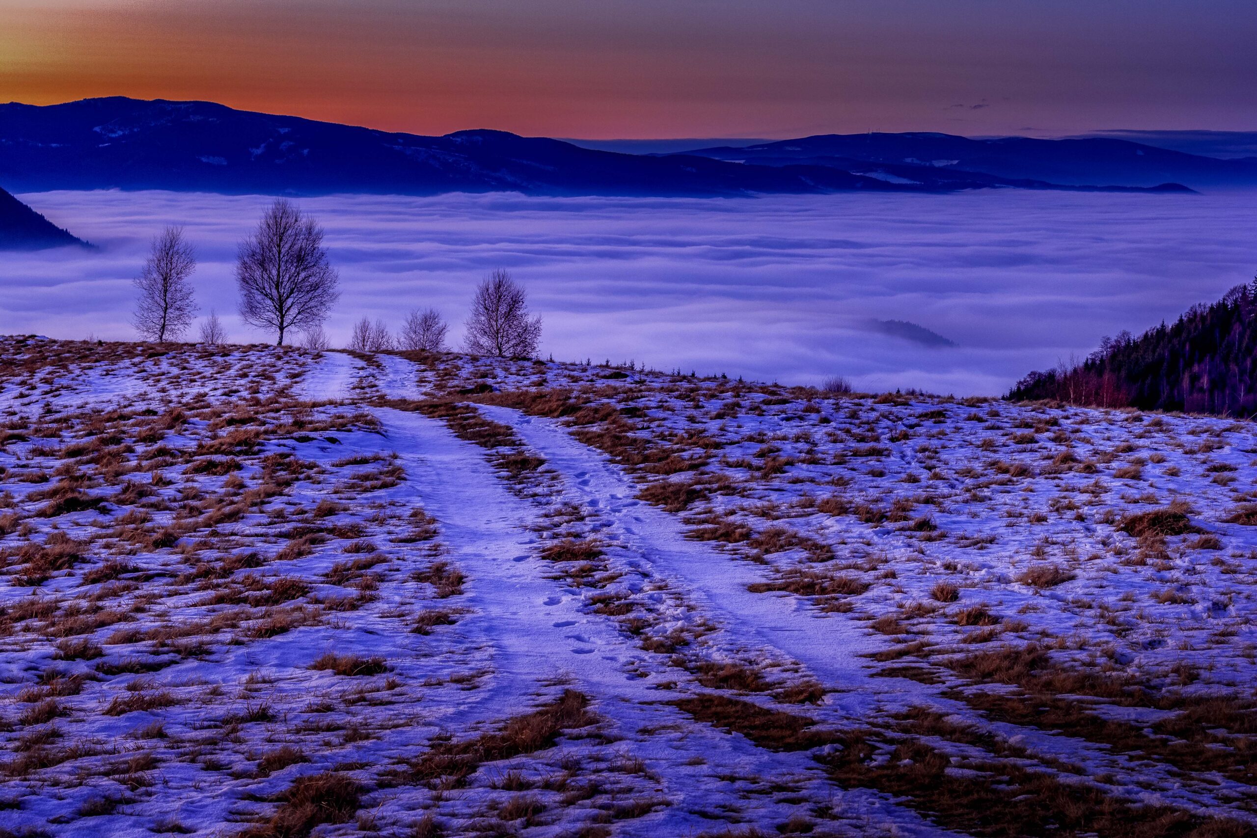 Winter view of Cecele Hill in Romania with snow-covered slopes, haze at the mountain base, and a vibrant sunset sky