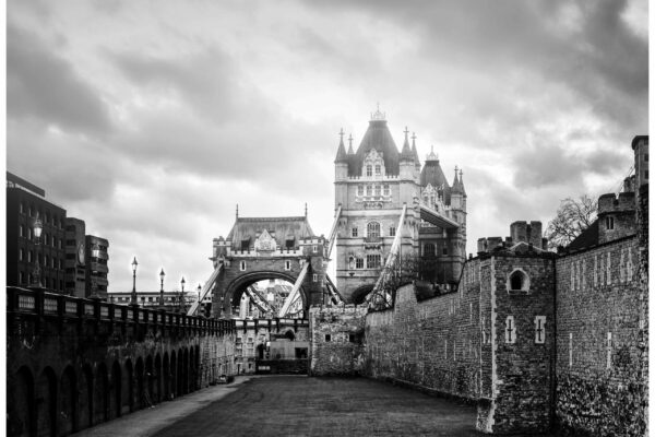 Black and white photograph of Tower Bridge viewed from the gardens near the Tower of London, with historic stone walls in the foreground
